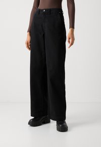 Črne široke hlače iz bombaža z gladko teksturo, z zapiranjem na gumb spredaj in stranskimi žepi. Obute z masivnimi črnimi škornji.