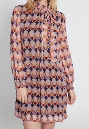 Femme portant une robe à manches longues et à motif avec un nœud noué au niveau de l'encolure dans des tons de violet, orange, rose et beige.