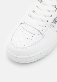 Sneaker bianca con pelle liscia, punta traforata per la ventilazione, lacci bianchi piatti e suola traforata. Presenta un marchio sul lato.