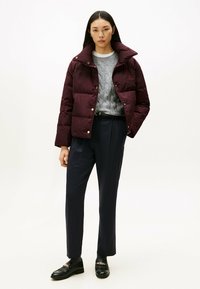 Burgundowa kurtka puchowa z wysokim kołnierzem i złotymi akcentami, w połączeniu z szarym swetrem w warkocze, granatowymi spodniami i czarnymi mokasynami.