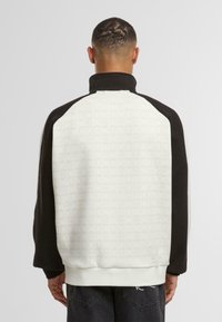 Pulover z barvnimi bloki ima belo hrbtno stran z diskretnim vzorcem, črne dolge rokave, visok ovratnik ter rebraste manšete in spodnji rob.