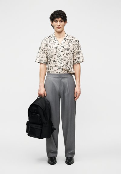 Νεαρός με σγουρά μαλλιά, που φοράει λευκό πουκάμισο με σχέδια και κοντά μανίκια, γκρι παντελόνι, μαύρα παπούτσια, κρατώντας ένα μαύρο σακίδιο σε ένα χέρι.