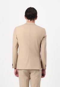Blazer beige con una vestibilità su misura, apertura posteriore singola e polsini a due bottoni. Texture liscia e design minimalista. Abbinato a pantaloni coordinati.