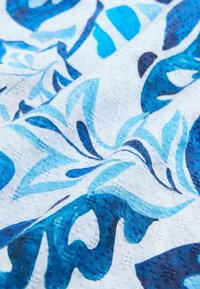 Blaues Stoffmuster mit floralen Motiven in hellen und dunklen Blautönen auf einem weißen Hintergrund, mit glatten Drapierungen und kunstvollen Details.