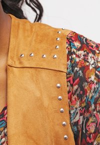 Hnědá semišová vesta se stříbrnými cvočky na ramenou, doplněná barevným vzorovaným topem s květinovými motivy na tmavém pozadí.