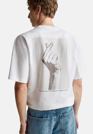 Witte katoenen t-shirt met een grafische print van een hand die een hartteken maakt. Korte mouwen en een relaxte pasvorm. Tekst onder de print.