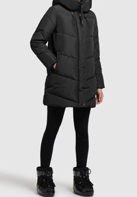 Černá péřová bunda s kapucí, se zipovým zapínáním, elastickými šňůrkami a velkým horizontálním prošíváním. V kombinaci s černými legínami a botami.