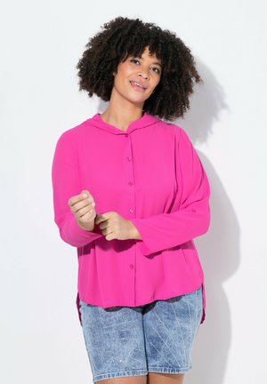 Maglietta a maniche lunghe, rosa brillante, con cappuccio e chiusura a bottoni, abbinata a pantaloncini in denim con motivo geometrico e decorazioni.