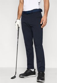 Μπλε σκούρα παντελόνια γκολφ με υφασμάτινη υφή, σε συνδυασμό με μαύρη ζώνη και παπούτσια. Ένα μπαστούνι γκολφ κρατείται στο αριστερό χέρι, ενώ στο δεξί υπάρχει άσπρο γάντι.