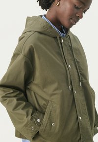 Jachetă cu glugă de culoare verde olive, realizată dintr-un material neted, având capse, șnururi și buzunare laterale. Designul este confortabil și practic.