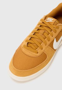 Nike tenisky v horčicovej žltej farbe s textúrovaným vrchným materiálom, bielym logom Swoosh a plochými šnúrkami. Zaoblený špička a gumová podrážka.