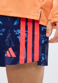 Κοντινό πλάνο ενός ατόμου που φοράει μπλε σορτς Adidas Adizero με έντονες κόκκινες ρίγες και πορτοκαλί μακρυμάνικη μπλούζα, με το χέρι χαλαρό στο πλάι.