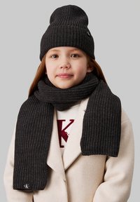 Beanie a coste grigio scuro e sciarpa abbinata realizzate in materiale morbido. La sciarpa è lunga e presenta un'etichetta dettaglio sul fondo.