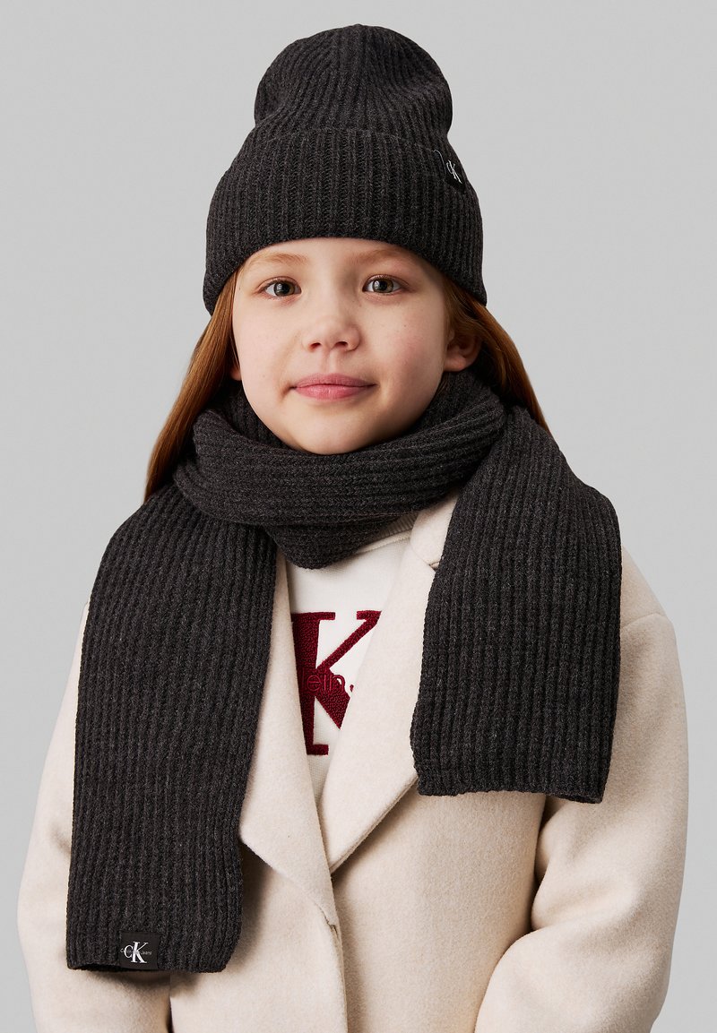 Beanie a coste grigio scuro e sciarpa abbinata realizzate in materiale morbido. La sciarpa è lunga e presenta un'etichetta dettaglio sul fondo.