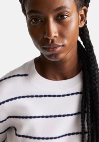 Biały sweter z granatowymi poziomymi paskami, okrągłym dekoltem i prążkowanym wykończeniem. Posiada teksturowany materiał i luźny krój.