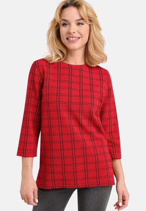 Rotes, kariertes Langarmshirt mit rundem Ausschnitt, aus strukturiertem Stoff mit schwarzem Gittermuster. Kombiniert mit grauer Hose.