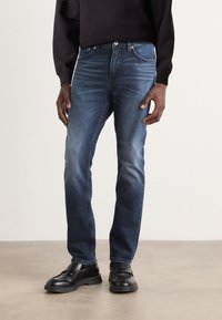 Jeans de mezclilla azul oscuro con un diseño ajustado, que presentan un estilo clásico de cinco bolsillos y detalles de desgaste sutiles, combinados con mocasines negros.