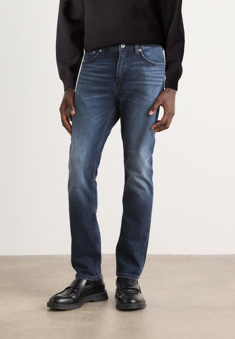 Jeans de mezclilla azul oscuro con un diseño ajustado, que presentan un estilo clásico de cinco bolsillos y detalles de desgaste sutiles, combinados con mocasines negros.