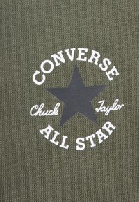 Olíva zöld szövet, texturált felülettel, fekete csillag logóval és fehér szöveggel, ami ezt olvasható: "CONVERSE Chuck Taylor ALL STAR."