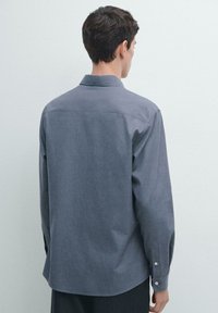 Šedá košile s dlouhým rukávem, límečkem, strukturovaným designem a jemnou texturou. Má zapínání na knoflíky na manžetách a zaoblený spodní lem, pohled zezadu.