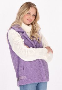 Lila, flauschige Weste mit hohem Kragen und Reißverschluss, über einem weißen, grob gestrickten Pullover geschichtet; verfügt über seitliche Taschen und einen verstellbaren Bund.