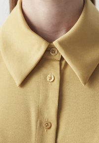 Gul skjorte med krave, der har et glat stof, med fire knapper foran og præcise syninger langs sømmene.