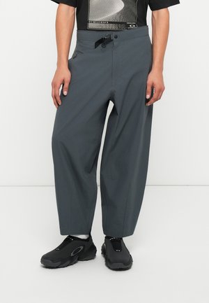 Sive, prostorne hlače z nastavljivim pasom, ki imajo stranske žepke in gladko tkanino. V kombinaciji z črnimi supergami na zavezovanje.