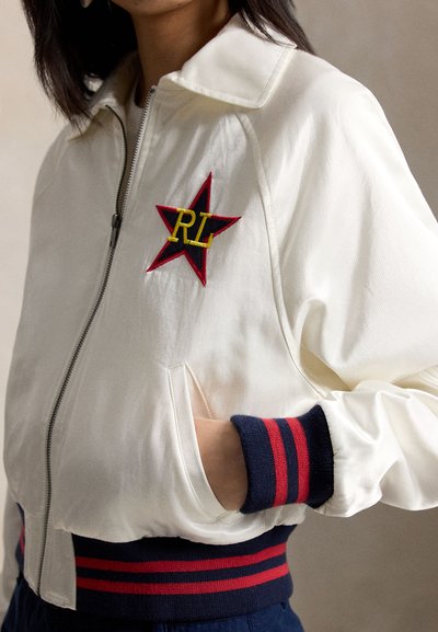 Žena nosící bílou bundu na zip s nárameníky a lemem s proužky v námořnické modři a červené barvě, a červenou nášivku ve tvaru hvězdy se žlutým nápisem "RL" na hrudi.