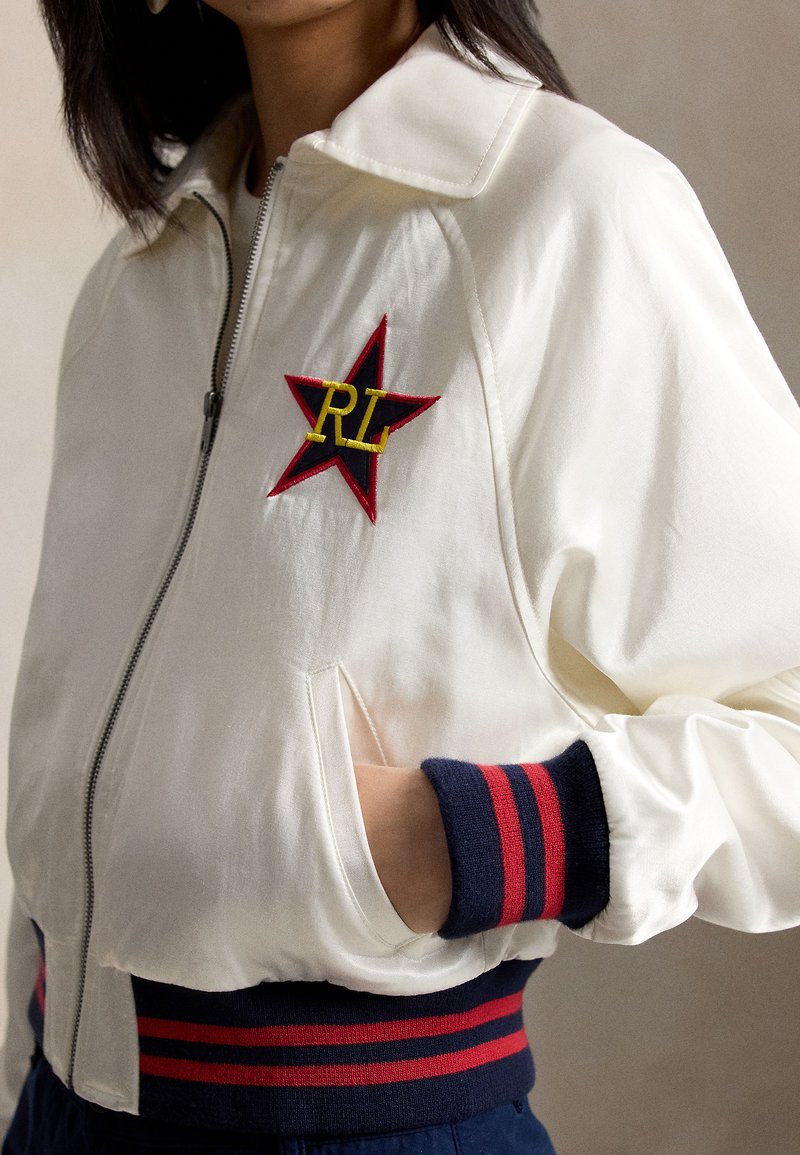Femme portant une veste blanche zippée avec des poignets et un ourlet rayés bleu marine et rouge, ainsi qu'un écusson étoile rouge avec les lettres jaunes "RL" sur la poitrine.