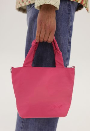 Hånd, der holder en lille lyserød håndtaske med polstrede håndtag, iført blå jeans og en langærmet skjorte med mønster.