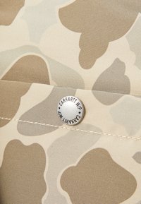 Poche en tissu camouflage avec un bouton en métal argenté, embossé avec "Carhartt WIP." Les couleurs incluent le beige et le marron clair, avec diverses formes organiques.