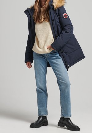 Femme portant un parka bleu marine avec capuche en fourrure, un pull beige, un jean bleu clair et des bottines noires, debout devant un fond gris uni.