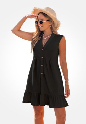 Mujer con vestido negro sin mangas con botones, sombrero de paja, gafas de sol y collares en capas, sombreando sus ojos con una mano.