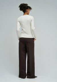 Donna con capelli ricci, indossante una maglietta bianca a maniche lunghe e pantaloni larghi scuri, in piedi con le mani in tasca, rivolta di spalle.