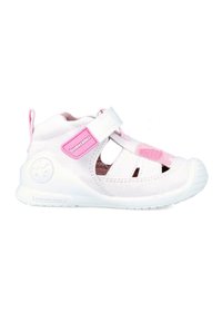 Zapato infantil de tela blanca con acentos rosas, diseño de ranuras abiertas, cinta de Velcro y suela de goma blanca. Presenta un logo en el lateral.