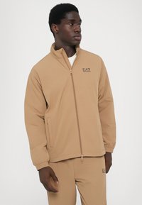 Tan zip-up džemperis, pagamintas iš minkštos medžiagos, su stovinčia apykakle, elastinėmis rankogaliais ir logo ant krūtinės. Pora su atitinkančiais kelnais.