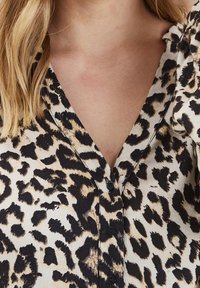 Leopardmönstrad blus med v-ringning, korta ärmar och svarta knappar. Tillverkad av lätta tyger med en slät textur och kontrasterande mönster.