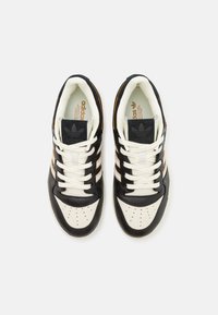 Svart och vit sportskor med tre guldstrimmor, en slät läderyta och en perforerad tå. Har vita snören och logotyp på tungan.