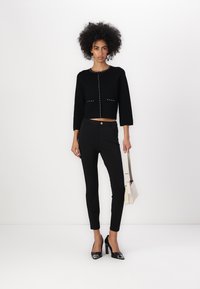 Suéter negro recortado con cuello redondo y tachuelas decorativas. Pantalones negros ajustados, zapatos de tacón altos negros y un bolso ligero.