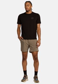 T-shirt atletico nero con logo, abbinato a pantaloni corti color khaki. Le caratteristiche includono una vestibilità rilassata e un tessuto traspirante. Indossato con calzini scuri e scarpe.