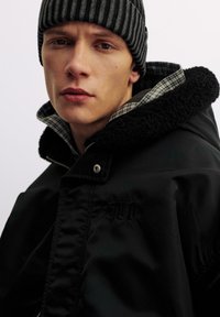 Schwarze Jacke mit einer strukturierten schwarzen Fleece-Kapuze, die ein kariertes Futter aufweist. Ein Knopfverschluss und ein gesticktes Logodetail sind erkennbar.