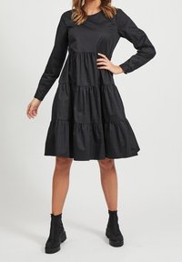 Schwarzes Langarmkleid mit gestuftem Rüschendesign, aus glattem Stoff gefertigt. Kombiniert mit schwarzen Knöchelstiefeln, hat einen runden Ausschnitt.