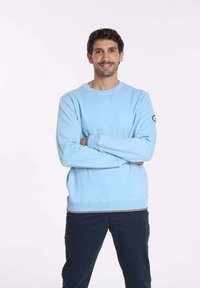 Pull bleu clair avec un col rond, des poignets côtelés et un ourlet avec une rayure contrastante. Le modèle se tient les bras croisés.