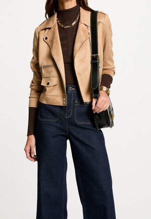 Femme portant une veste en daim beige, un pull à col roulé marron foncé, un jean large foncé, un collier en or, des bracelets, et portant un sac à épaule noir.