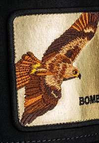 Izšūts plāksteris, kas attēlo lidojošu ērglīti brūnās un zeltainās krāsās uz teksturēta zelta fona, apmalots ar melnu audumu un tekstu "BOMB."