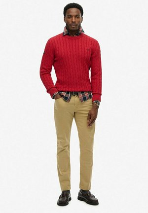 Pull en maille rouge avec une chemise à carreaux en dessous, associé à un chino beige et des chaussures en cuir noir. Présente un motif texturé sur le pull.