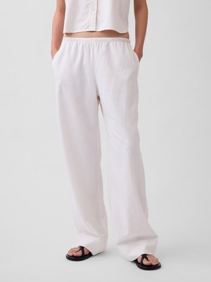 Personne portant un pantalon ample en lin blanc avec une taille élastique, un haut blanc court sans manches, et des sandales noires, debout devant un fond uni.