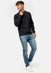 Sudadera negra con logo texturizado, combinada con jeans desgastados de color azul claro y zapatillas blancas. El modelo está en una pose relajada.