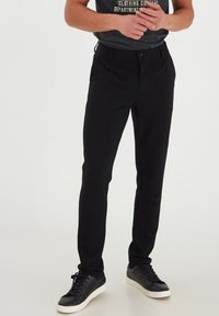 Schwarze Hose mit schmalem Schnitt, aus einem glatten Stoff. Mit Knopfverschluss und Seitentaschen, kombiniert mit schwarzen Sneakers.