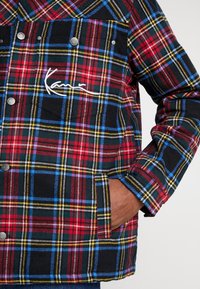 Rutigt skjorta med röda, svarta och gula färger samt blå detaljer. Framficka med tryckknappar och broderad logotyp på bröstet.